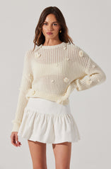A woman is wearing a cream-colored, loose-knit sweater with small 3D floral details scattered across it. She pairs it with a white, high-waisted, ruffled mini skirt.