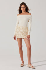 A woman is wearing an off-the-shoulder, long-sleeve white top paired with a cream-colored textured mini skirt. She has on white woven flats and gold hoop earrings.