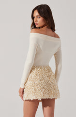 A woman is wearing an off-the-shoulder, long-sleeve, cream-colored top paired with a high-waisted, textured cream-colored skirt that features a floral or ruffled pattern.