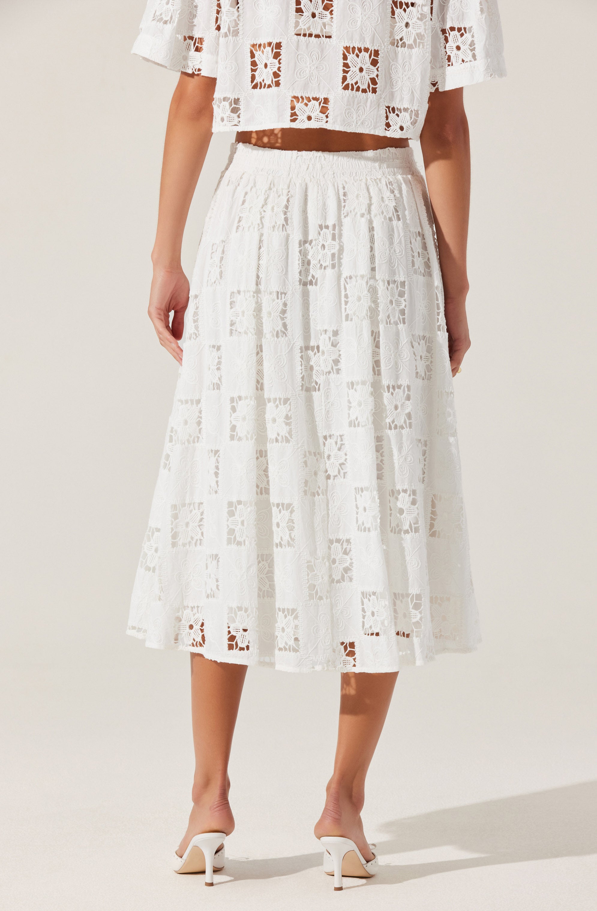 A woman is wearing a white, semi-sheer lace skirt with a patchwork pattern of squares and floral designs. The skirt has a high waist with an elastic band and falls to mid-calf length. She is also wearing white high-heeled shoes with ankle straps. The top she is wearing is a matching white lace off-the-shoulder crop top with a similar patchwork pattern.