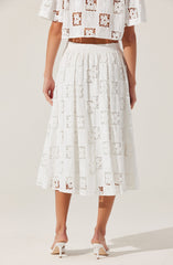 A woman is wearing a white, semi-sheer lace skirt with a patchwork pattern of squares and floral designs. The skirt has a high waist with an elastic band and falls to mid-calf length. She is also wearing white high-heeled shoes with ankle straps. The top she is wearing is a matching white lace off-the-shoulder crop top with a similar patchwork pattern.
