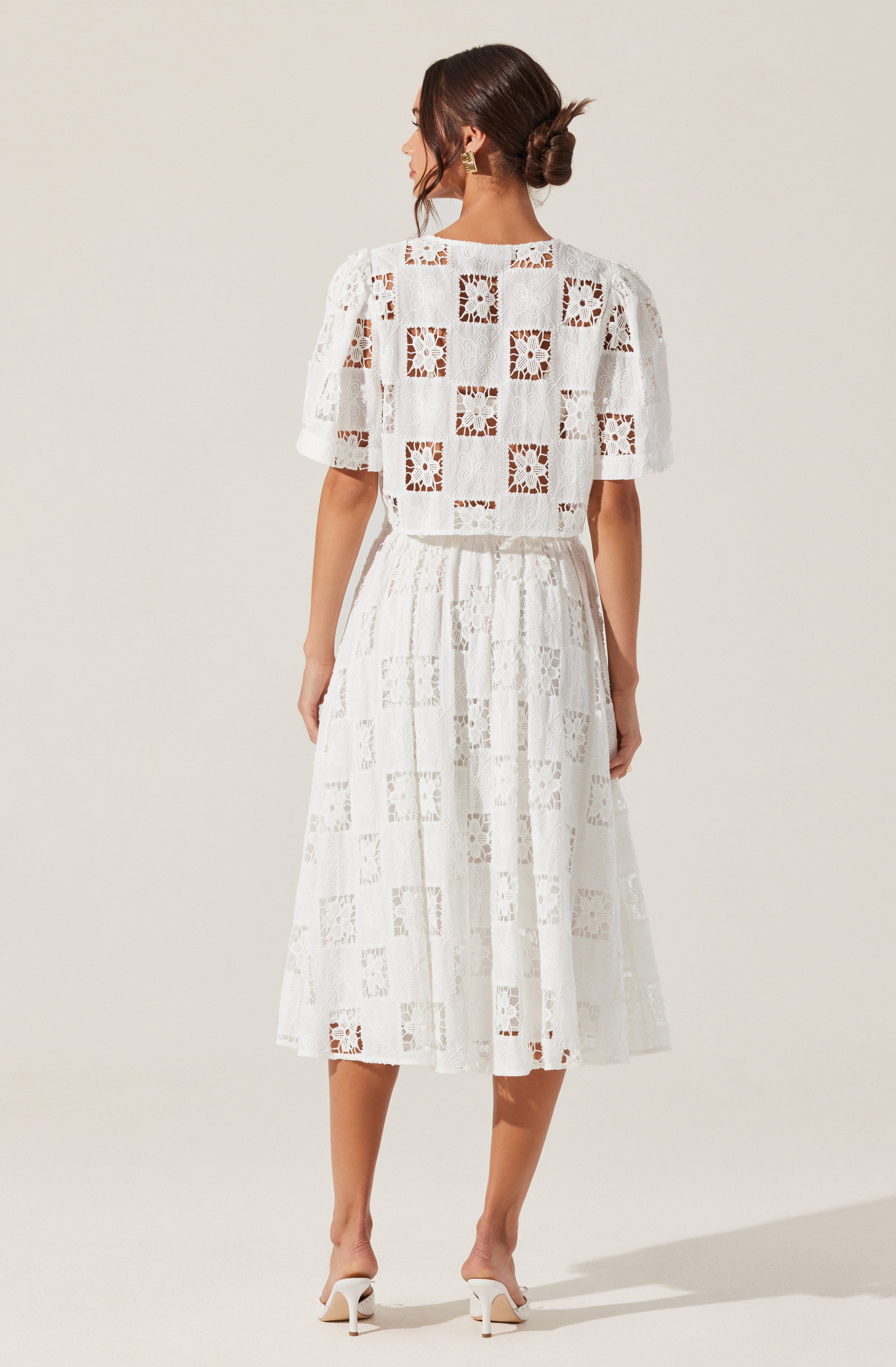 A woman is wearing a white, lace dress with a pattern of square and floral cutouts. The dress has a loose-fitting top with short sleeves and a high neckline, and a gathered, mid-length skirt that falls below the knees. She is also wearing white high-heeled shoes.
