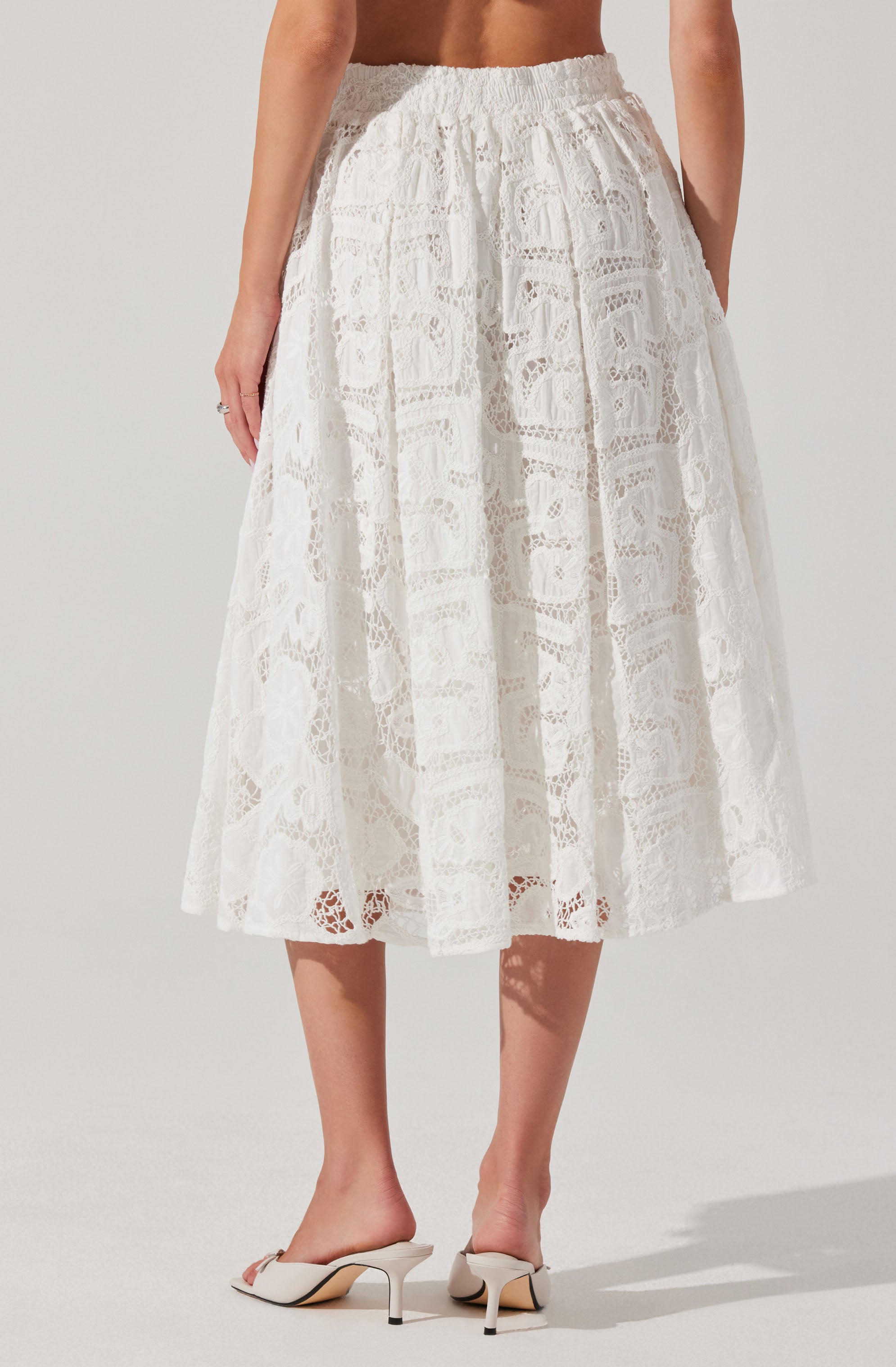 The image shows a woman wearing a white lace midi skirt with an elastic waistband and a drawstring tie at the waist. The skirt features intricate lace patterns with floral and geometric designs, creating a semi-sheer effect. She is also wearing white high-heeled sandals with an open toe design.