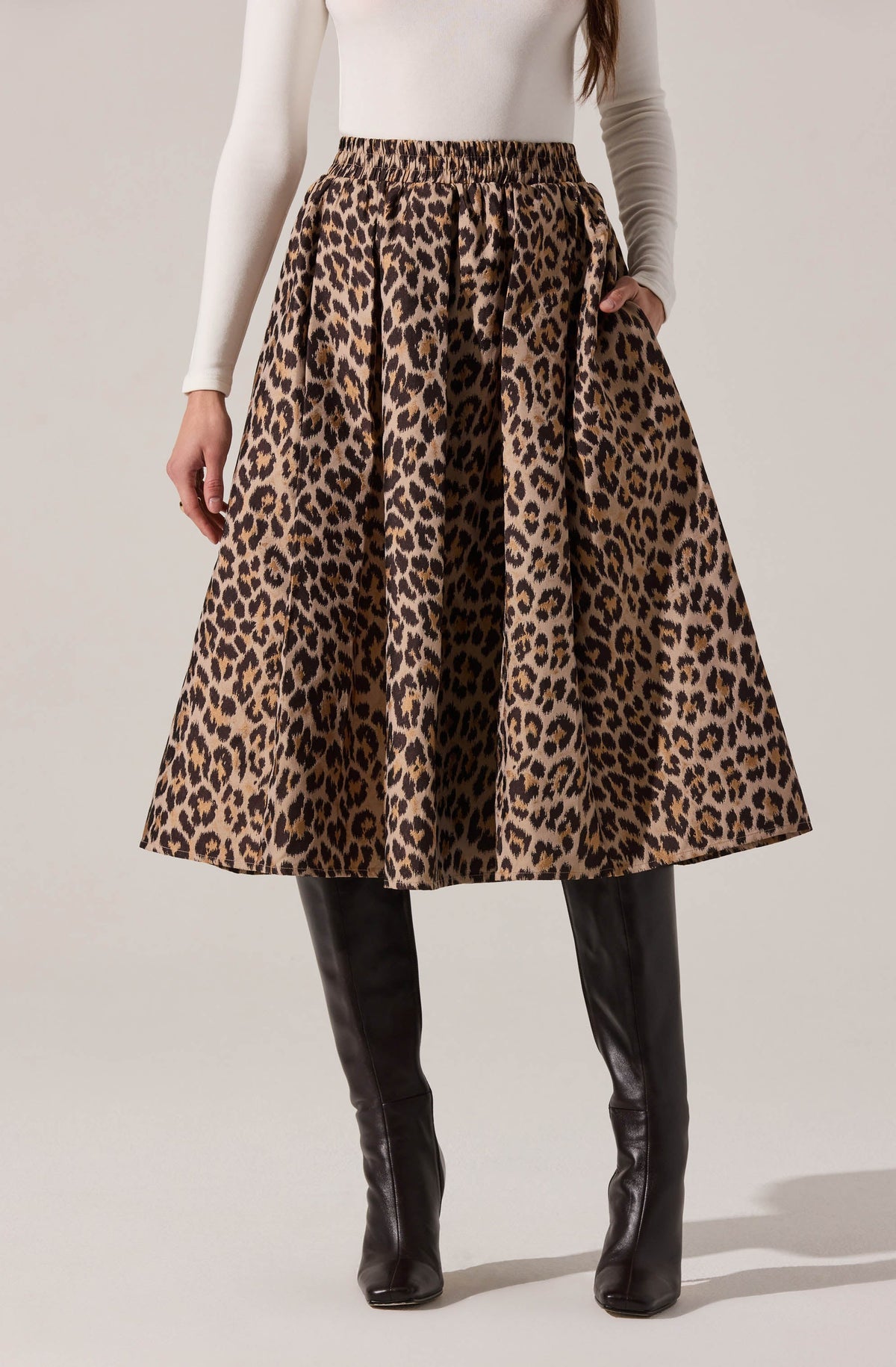A woman is wearing a high-waisted, full-length skirt with a bold leopard print pattern in shades of brown and black. She pairs it with black, knee-high leather boots and a fitted, long-sleeved white top.