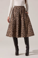 A woman is wearing a high-waisted, full-length skirt with a bold leopard print pattern in shades of brown and black. She pairs it with black, knee-high leather boots and a fitted, long-sleeved white top.