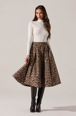 A woman is wearing a fitted white long-sleeve turtleneck top paired with a high-waisted, ankle-length leopard print skirt that has a voluminous, flowing silhouette. She completes the outfit with black knee-high boots.