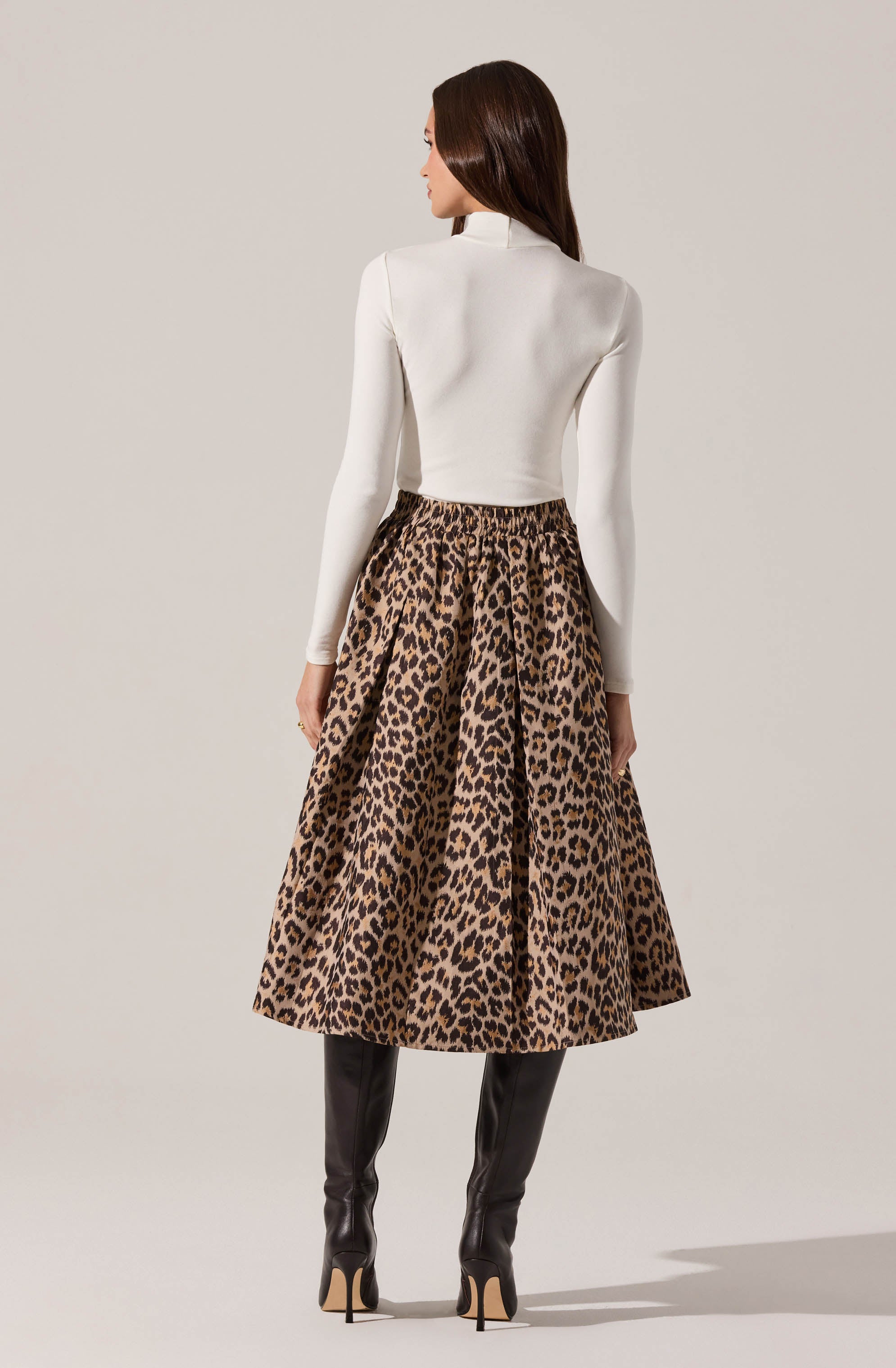 A woman is wearing a fitted white long-sleeve top and a high-waisted, full-length leopard print skirt with a voluminous silhouette. She also has on tall, black leather knee-high boots with high heels.