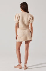 A woman is wearing a beige, textured two-piece outfit consisting of a short-sleeved, puff-sleeve sweater with a fitted waistband, and a matching short skirt. The sweater and skirt are decorated with small, raised pearl-like embellishments. She is also wearing beige ballet flats with ankle ribbons tied in bows.