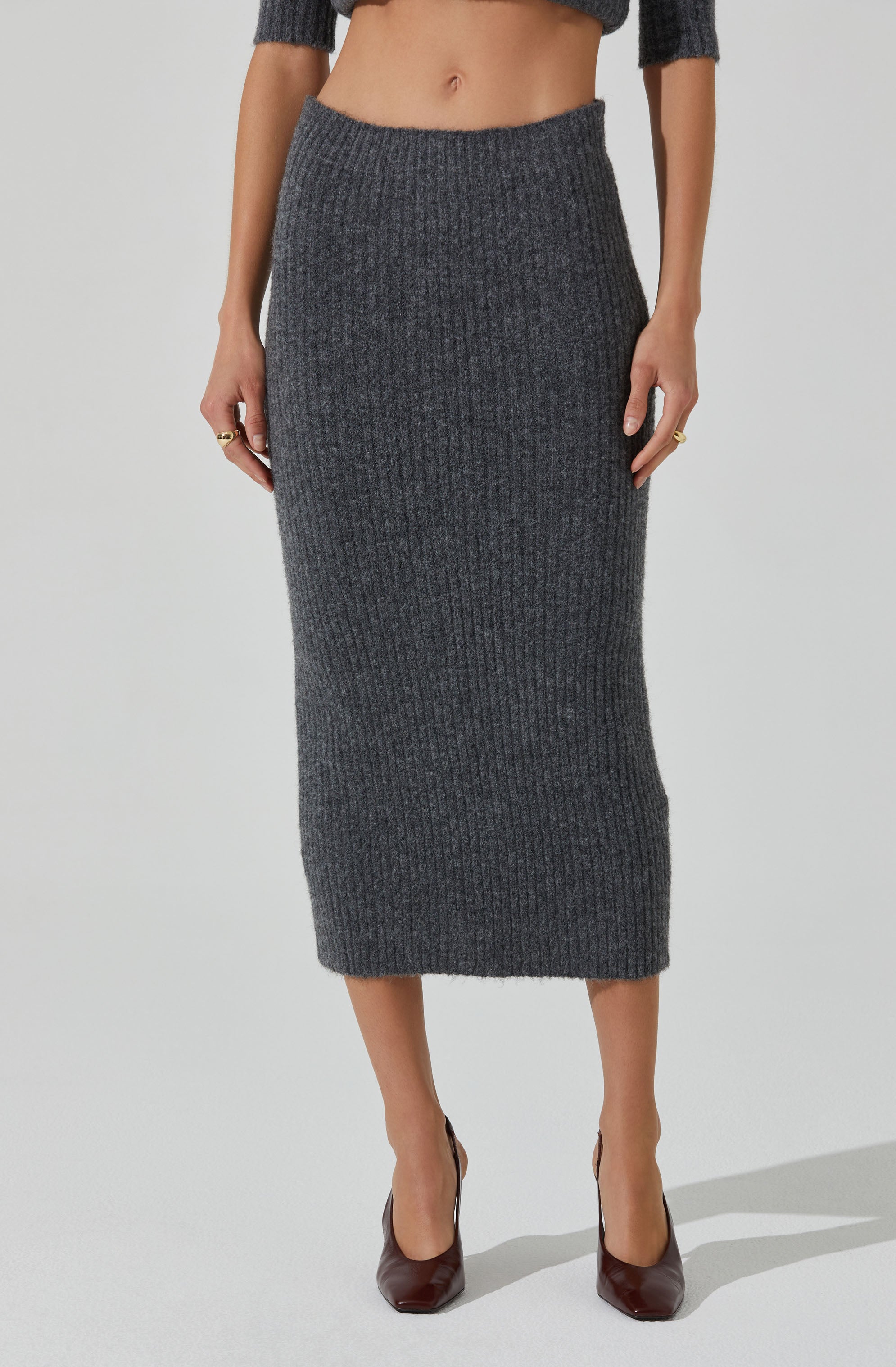 A woman is wearing a dark gray, high-waisted, ribbed knit skirt that extends to mid-calf length. She is also wearing a matching dark gray, cropped, long-sleeve sweater.