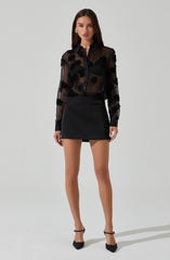 A woman is wearing a sheer black blouse with floral embroidery details, paired with a high-waisted black mini skirt, and black pointed-toe high heels with ankle straps.