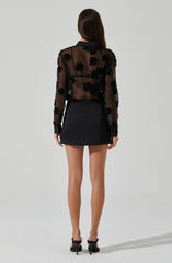 A woman is wearing a black sheer long-sleeve blouse with floral appliqué details, paired with a black mini skirt. She is also wearing black high-heeled shoes.
