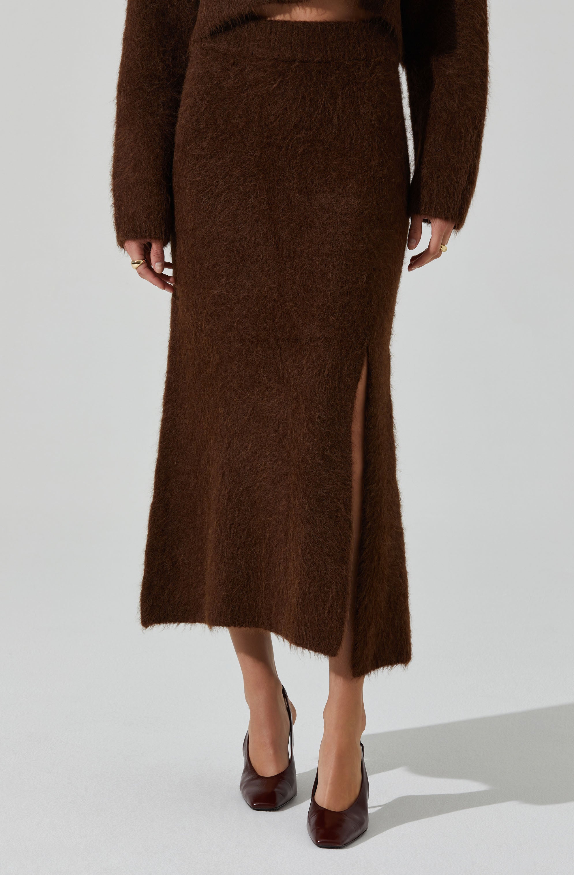 A woman is wearing a matching brown fuzzy knit sweater and skirt set. The skirt has a high slit on one side, revealing her leg and a pair of dark brown high-heeled shoes.