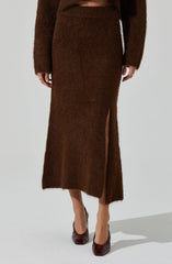 A woman is wearing a matching brown fuzzy knit sweater and skirt set. The skirt has a high slit on one side, revealing her leg and a pair of dark brown high-heeled shoes.