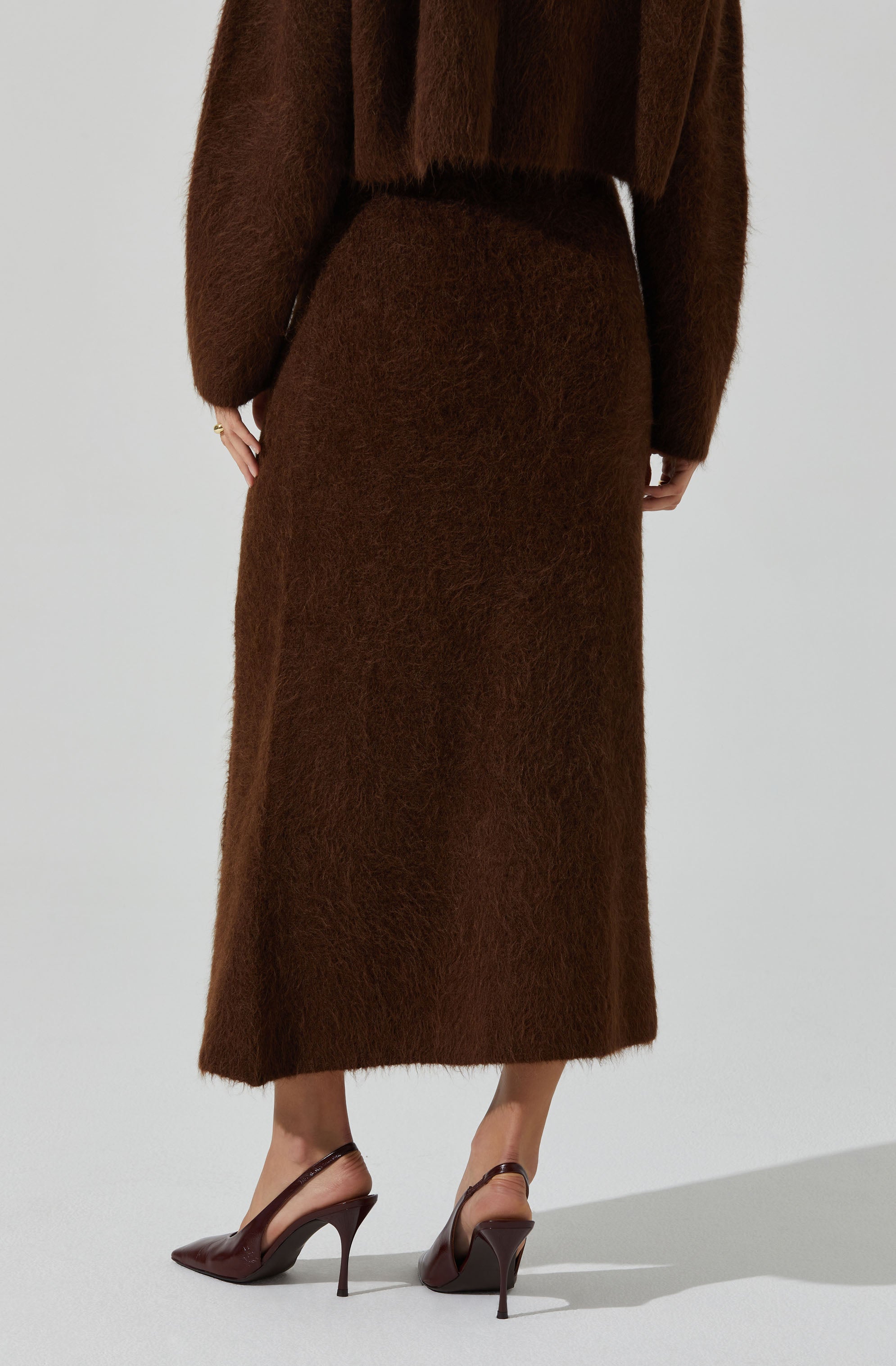 A person is wearing a long, brown, fuzzy skirt that reaches the ankles. They are also dressed in a matching brown, fuzzy top with long sleeves. The outfit appears cozy and warm, with the top slightly loose-fitting. The person is wearing brown high-heeled shoes with pointed toes and ankle straps.