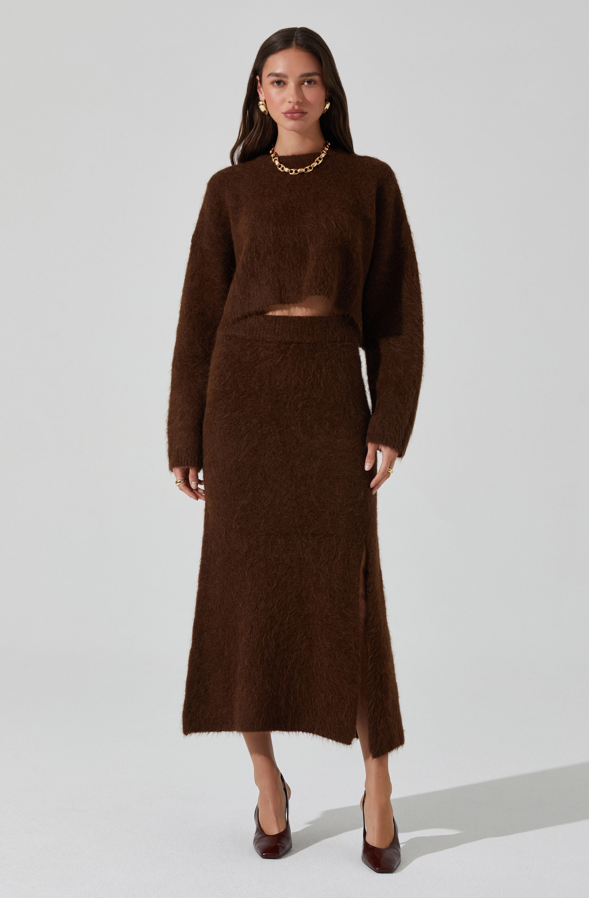 A woman is wearing a matching brown fuzzy knit set consisting of a long-sleeved cropped sweater and a high-waisted, ankle-length skirt with a side slit. She is accessorized with gold jewelry, including hoop earrings and a chunky chain necklace, and is wearing dark brown heels.