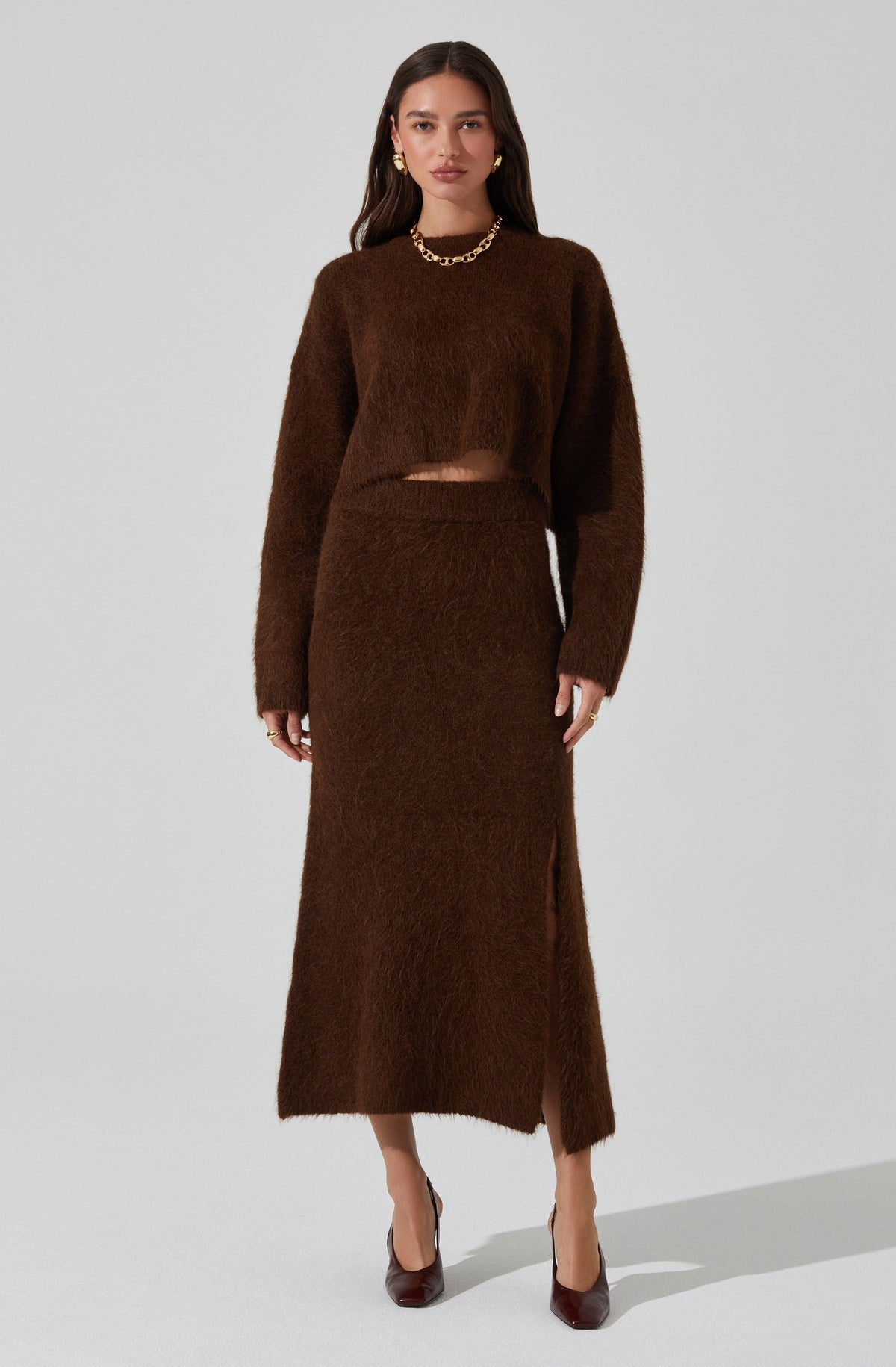 A woman is wearing a matching brown fuzzy knit set consisting of a long-sleeved cropped sweater and a high-waisted, ankle-length skirt with a side slit. She is accessorized with gold jewelry, including hoop earrings and a chunky chain necklace, and is wearing dark brown heels.