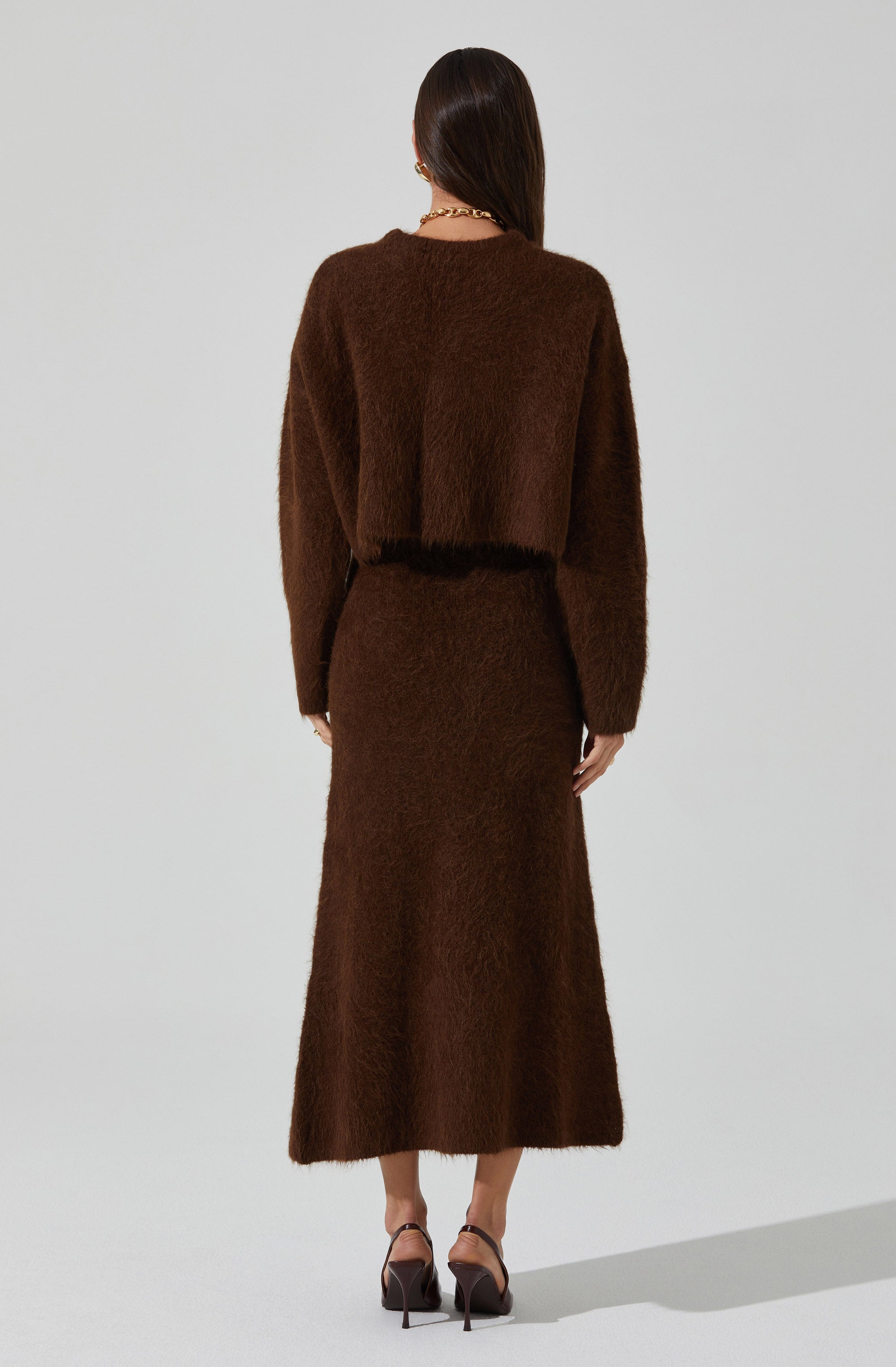 A woman is wearing a matching brown fuzzy knit outfit consisting of a cropped sweater with long sleeves and a high-waisted, ankle-length skirt. She is accessorized with gold jewelry, including earrings and a chain necklace, and is wearing black strappy high-heeled sandals.