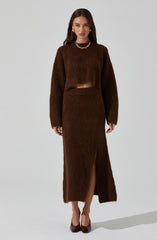 A woman is wearing a matching set of a brown fuzzy long-sleeve top and a high-waisted skirt with a front slit. She accessorizes with a chunky gold chain necklace, gold earrings, and rings, and is wearing dark brown high heels.