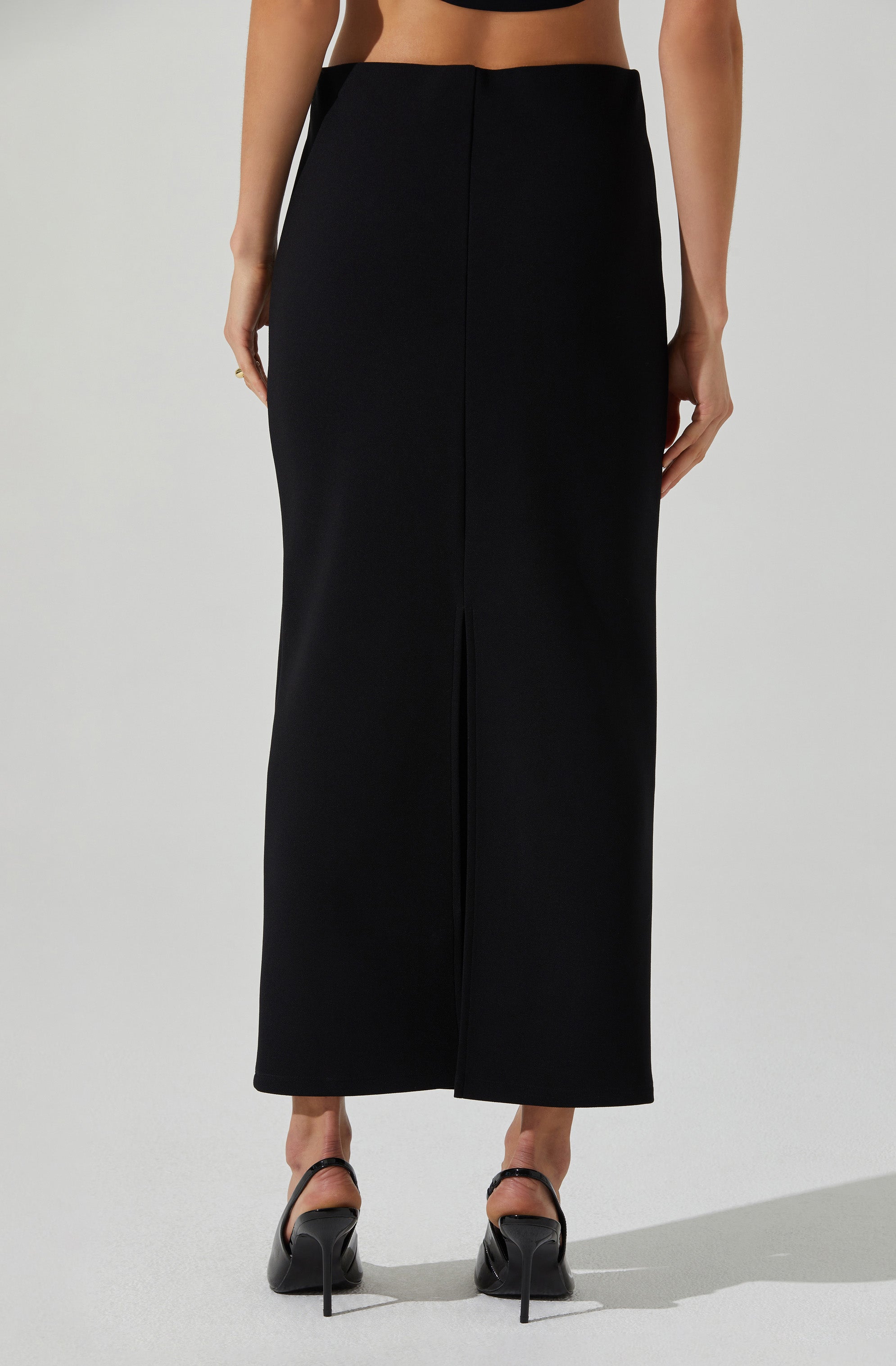 A woman is wearing a high-waisted, wide-leg black skirt that extends to her ankles. She has paired it with black high-heeled shoes with ankle straps. The skirt has a central front slit and is made of a smooth, slightly shiny fabric.