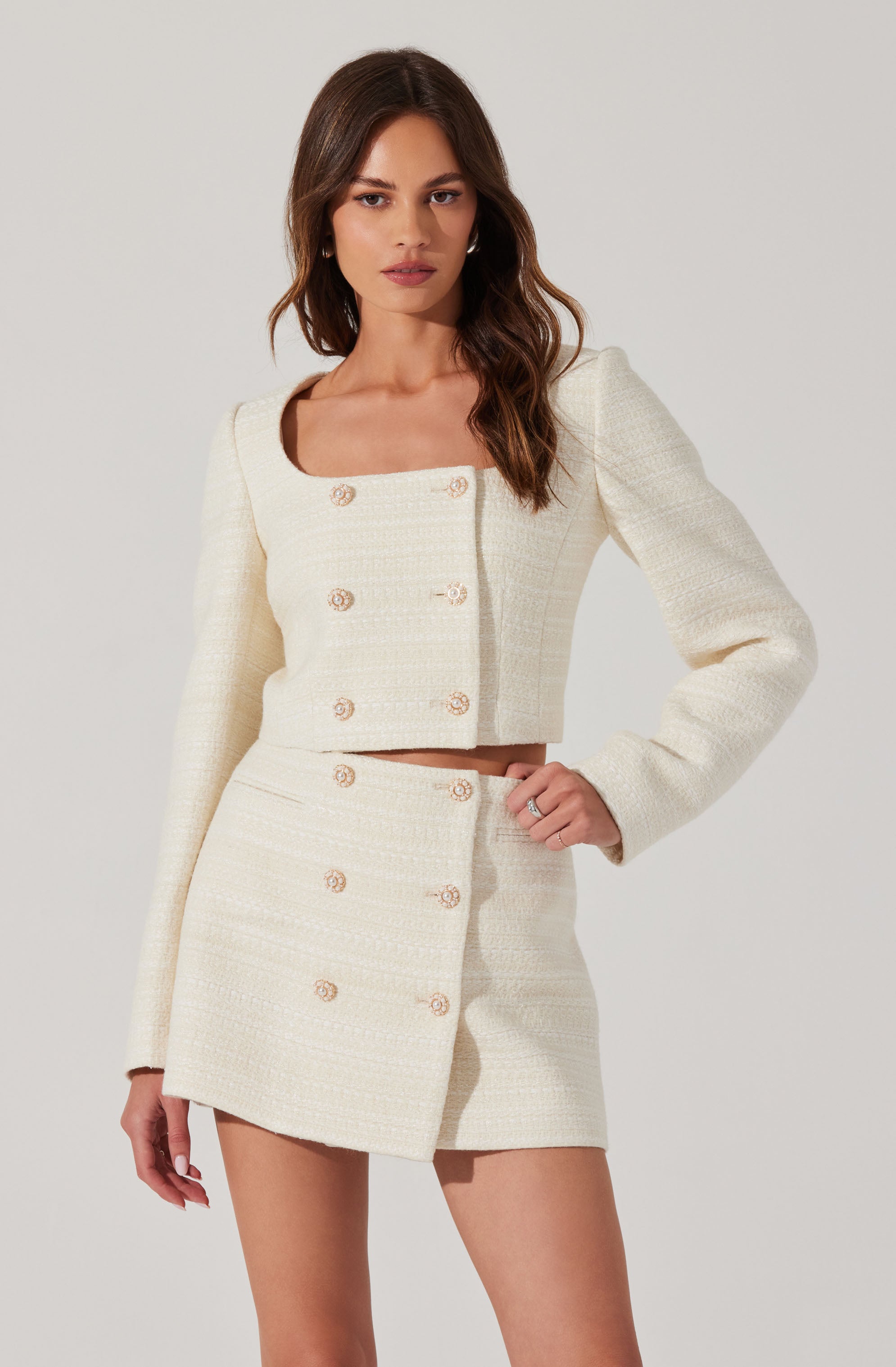 A woman is wearing a two-piece outfit made of cream-colored, textured fabric. The top is a cropped, long-sleeved jacket with a square neckline, featuring a double row of gold buttons down the front. The matching high-waisted skirt is short and has two rows of gold buttons running vertically along the front.