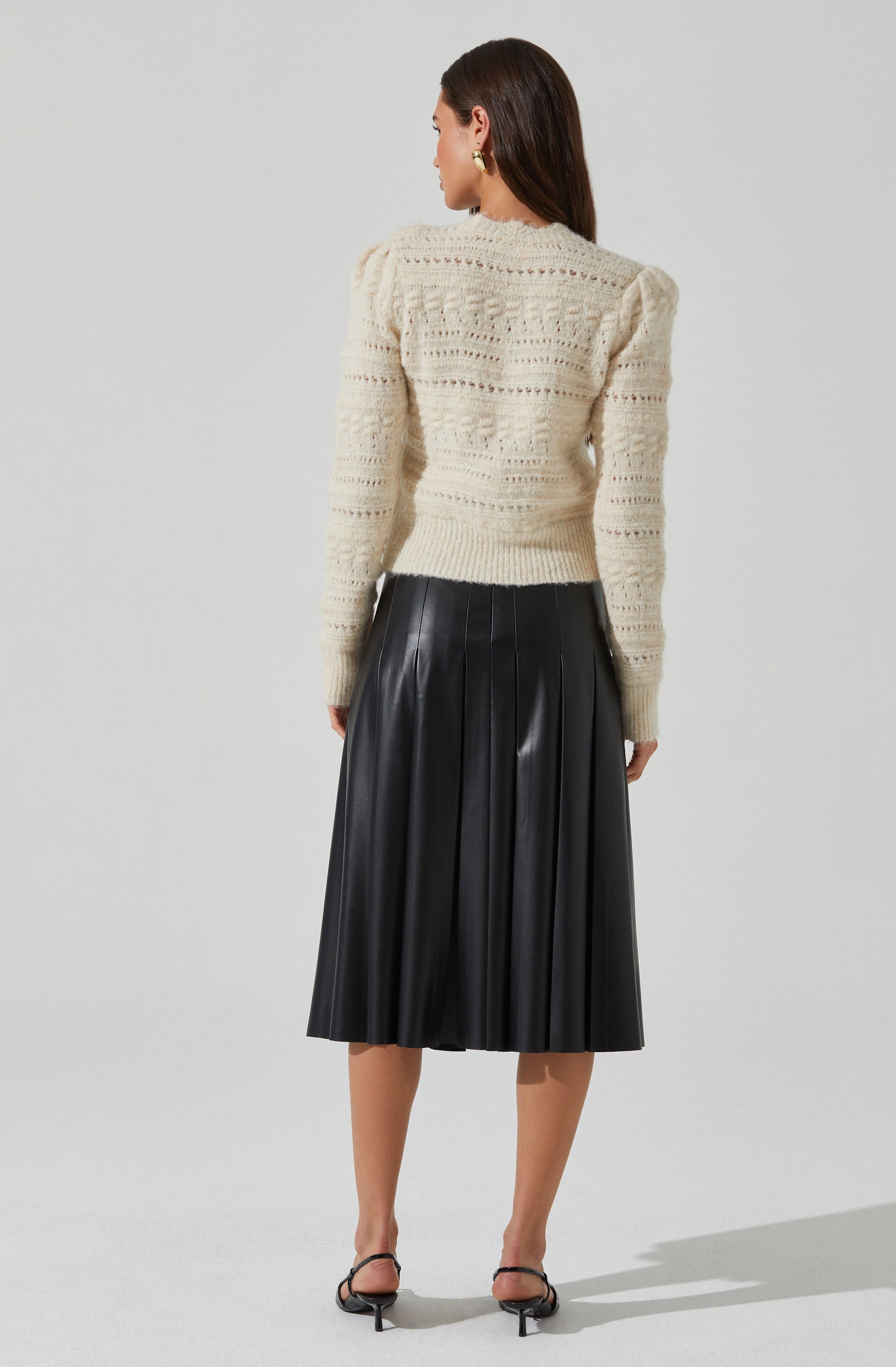 A woman is wearing a cream-colored, long-sleeved, knitted sweater with a textured pattern. The sweater has a fitted silhouette and slightly puffed shoulders. She pairs it with a black, high-waisted, pleated midi skirt made of a shiny material, and black open-toed heels with ankle straps.
