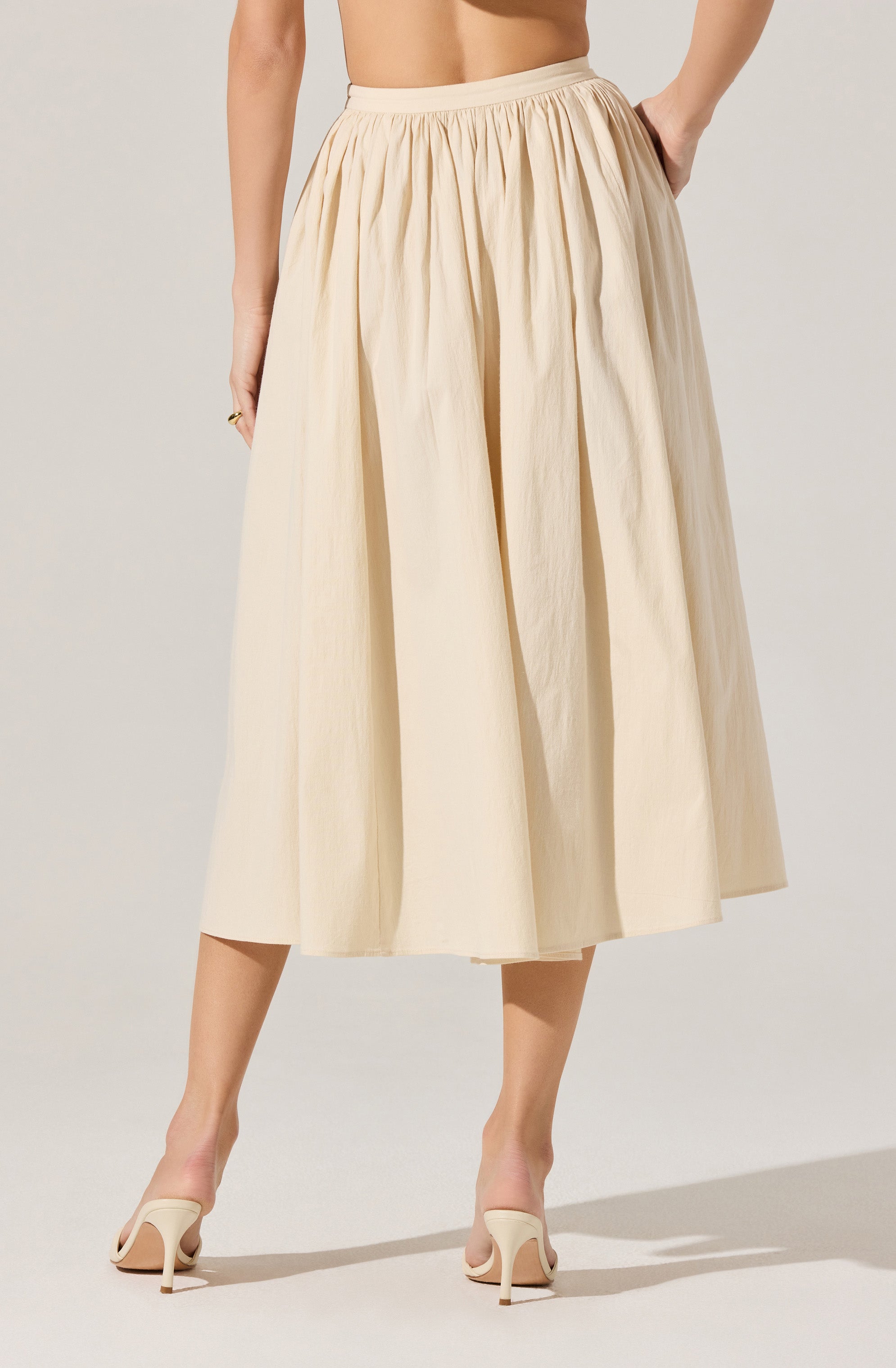 A woman is wearing a cream-colored, ankle-length skirt with a gathered waistband and wide pleats. She pairs the skirt with nude high-heeled shoes. Her hand rests on her hip, showing part of her torso.
