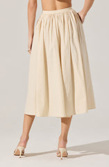 A woman is wearing a cream-colored, ankle-length skirt with a gathered waistband and wide pleats. She pairs the skirt with nude high-heeled shoes. Her hand rests on her hip, showing part of her torso.