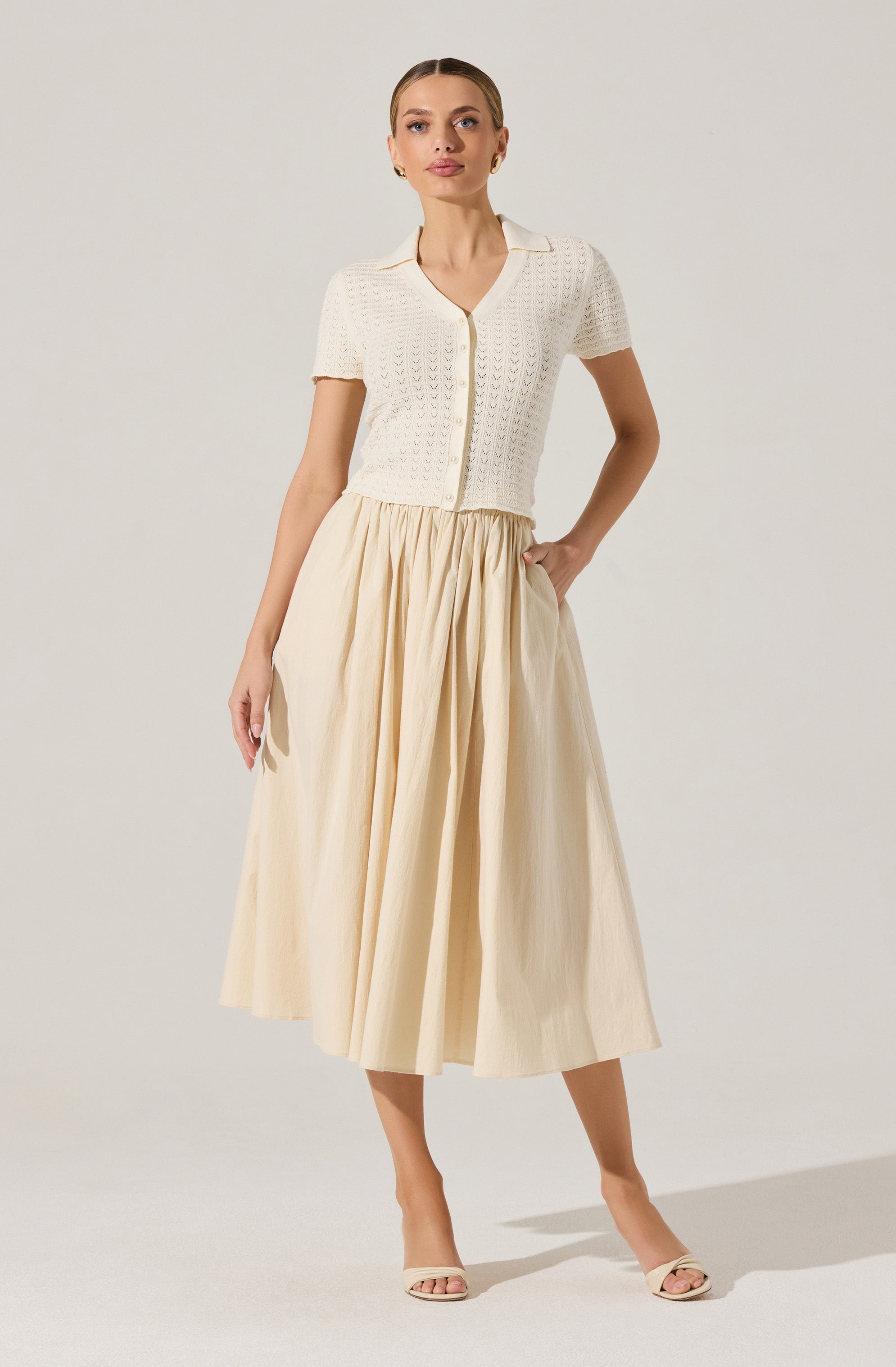 A woman is wearing a short-sleeved, cream-colored knitted cardigan with a V-neck and button closure in the front. The cardigan has a textured, openwork pattern. She pairs it with a high-waisted, long, pleated skirt in a light, neutral color that reaches mid-calf. She completes the outfit with light-colored open-toe heeled sandals.