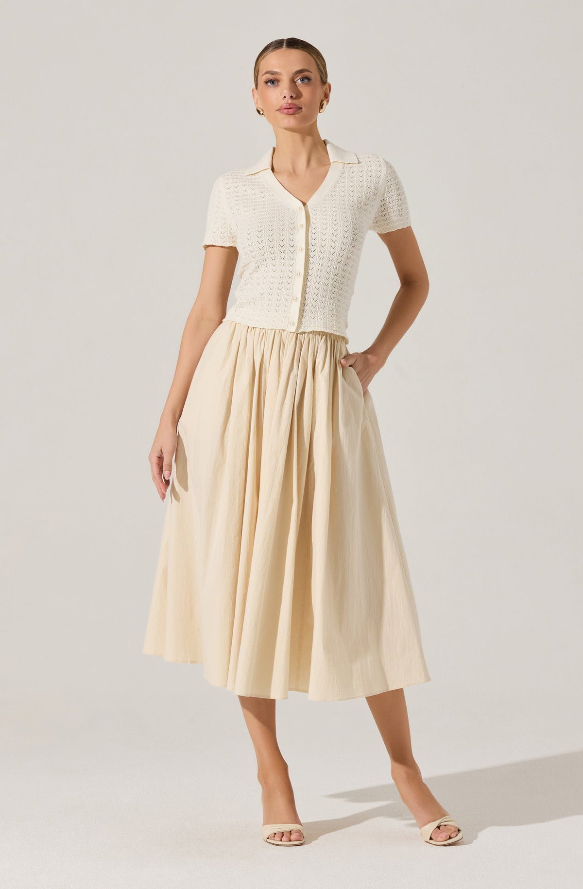 A woman is wearing a short-sleeved, cream-colored knitted cardigan with a V-neck and button closure in the front. The cardigan has a textured, openwork pattern. She pairs it with a high-waisted, long, pleated skirt in a light, neutral color that reaches mid-calf. She completes the outfit with light-colored open-toe heeled sandals.