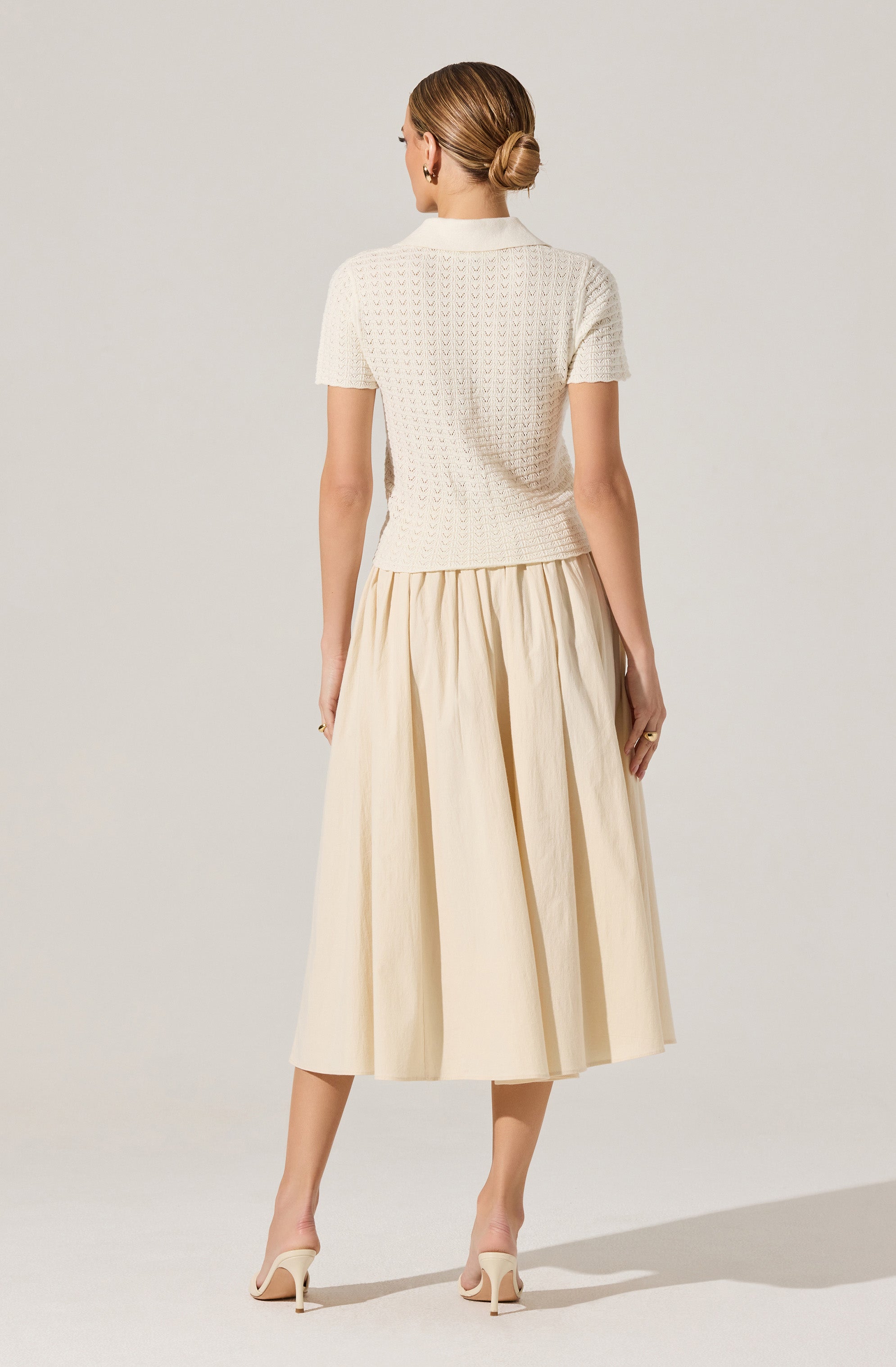 A woman is wearing a short-sleeved, light-colored, textured knit top with a collar. She pairs it with a high-waisted, full-length, beige skirt that has a gathered, pleated design. She is also wearing light-colored high-heeled shoes.