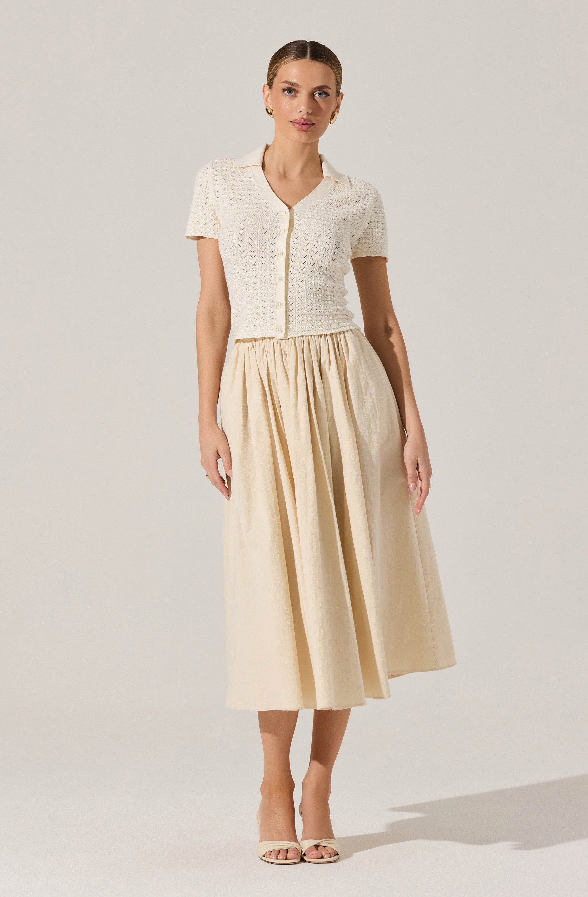 A woman is wearing a short-sleeved, open-knit, cream-colored cardigan with a collar and buttons down the front, paired with a high-waisted, beige, pleated midi skirt. She has on light-colored open-toe heels.