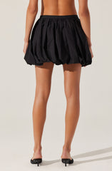 A person is wearing a black, voluminous, bubble-style skirt with a gathered, elastic waistband. The skirt has a shiny, slightly crinkled texture and a loose fit that creates a rounded silhouette. The individual is also wearing black high-heeled shoes with open toes and thin ankle straps.
