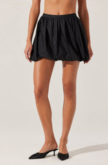 A person is wearing a black, voluminous, puffed skirt with a gathered waistband. They are also wearing black high-heeled shoes with pointed toes and decorative bows on the front.