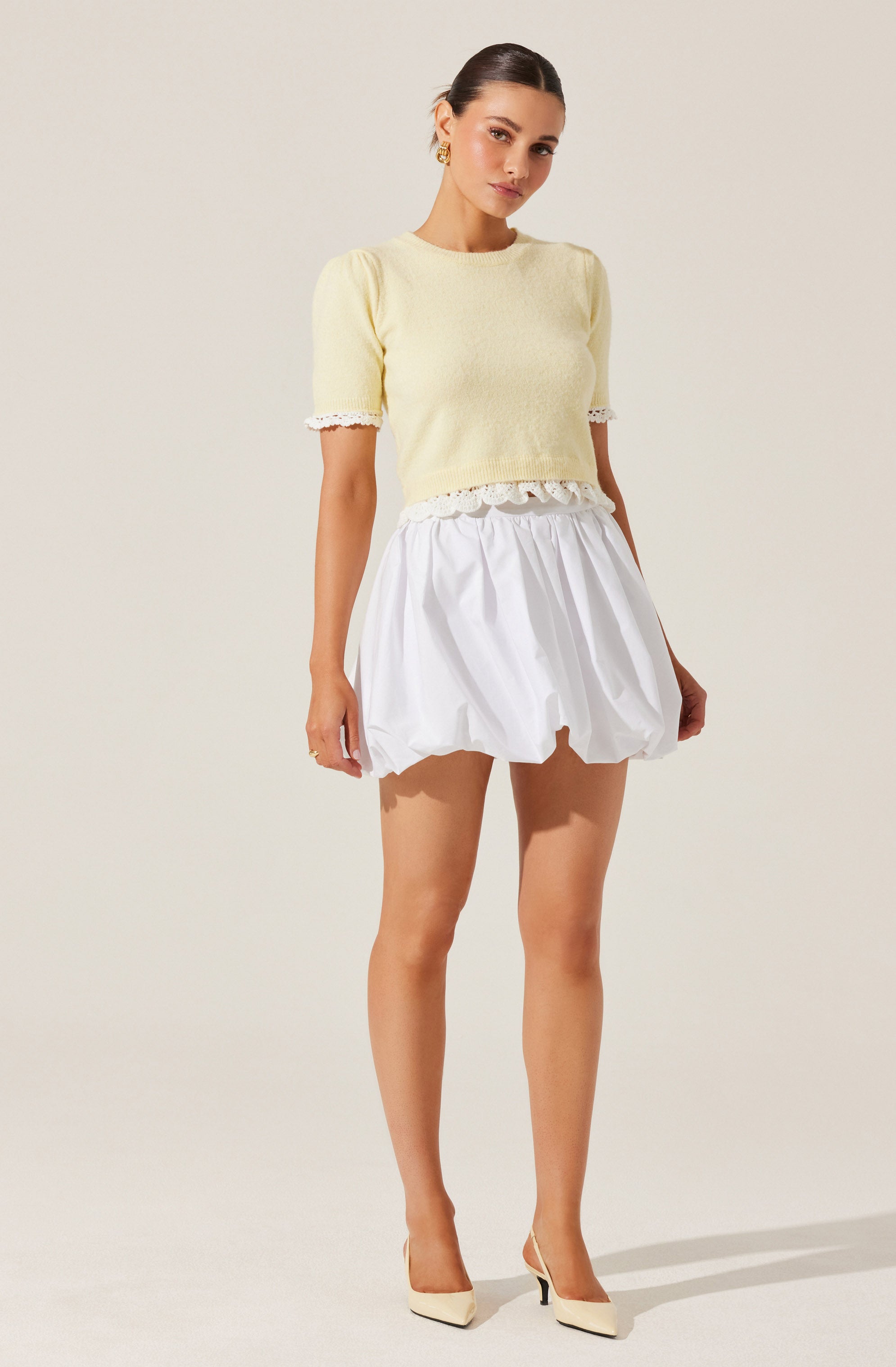 A woman is wearing a light yellow, short-sleeved, cropped knit top with lace trim on the sleeves and hem. She pairs it with a white, puffed, knee-length skirt with an elastic waistband. She completes the outfit with beige high-heeled shoes.