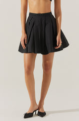 A woman is wearing a high-waisted black skirt with pleats and vertical piping details. She is also wearing black pointed-toe high heels with a bow detail. Her hands are relaxed by her sides, with one hand showing a gold ring.