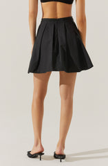 A woman is wearing a high-waisted, black, pleated skirt made of a shiny, lightweight fabric. The skirt has a waistband with belt loops and features sharp pleats, giving it a structured look. She is also wearing black open-toe heels with thin straps and a small heel. The outfit is shown against a plain, neutral background.