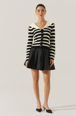 A woman is wearing a black and white striped cardigan with gold buttons, featuring a wide collar that drapes over her shoulders. She pairs the cardigan with a black pleated skirt and black high-heeled shoes with bows on the front.