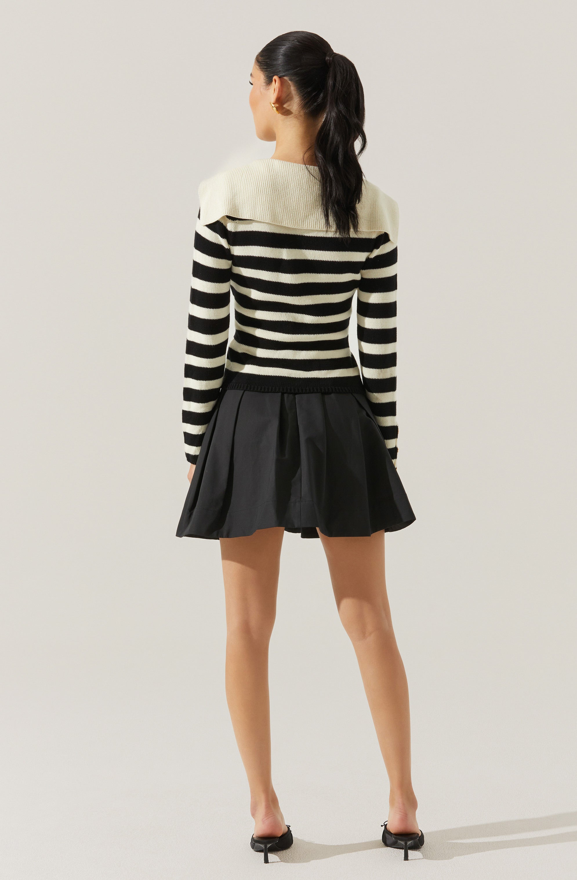 A woman is wearing a long-sleeved, crew-neck sweater with horizontal black and white stripes and a large white shoulder yoke. She pairs the sweater with a black pleated mini skirt and black open-toe heels with ankle straps.