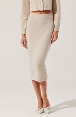 A woman is wearing a beige, knitted, long-sleeve cardigan with buttons, a matching beige, knitted midi skirt with a wide waistband, and white high-heeled shoes with a bow detail.