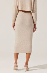 A woman is wearing a beige, knit, long-sleeve sweater and a matching beige knit midi skirt. She is also wearing white high-heeled sandals.