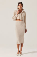 A woman is wearing a cream-colored, cropped, button-up sweater with a collar, paired with a matching high-waisted, mid-length knit skirt. She is also wearing light-colored, pointed-toe heels with ankle straps.