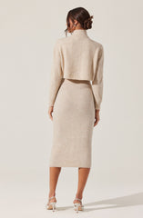 A woman is wearing a beige, knitted two-piece outfit consisting of a cropped turtleneck sweater with long sleeves and a matching high-waisted, midi-length skirt. She has her hair styled in a low bun and is wearing white high-heeled shoes.