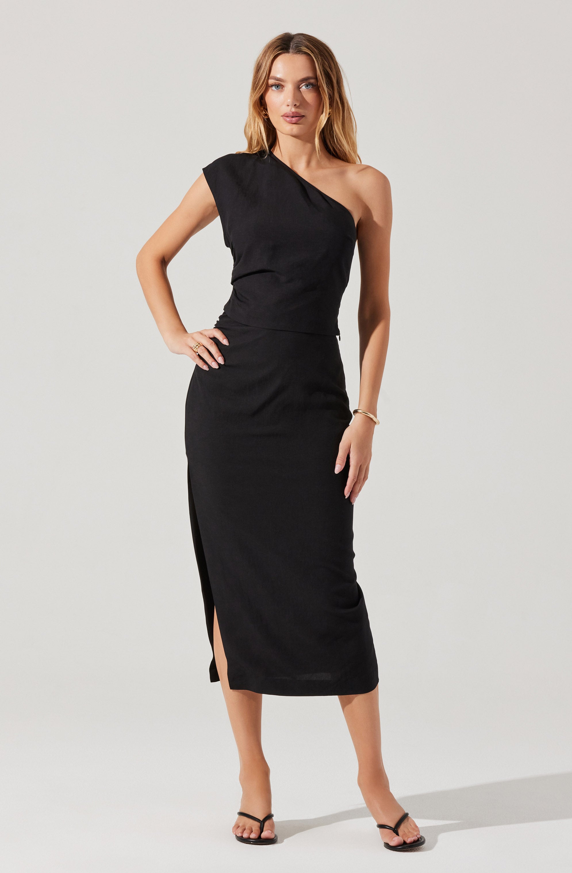 A woman is wearing a black dress with a one-shoulder design. The dress is fitted at the waist and extends down to mid-calf length, featuring a slit on the left side. She is also wearing black heeled sandals and a simple bracelet on her right wrist.