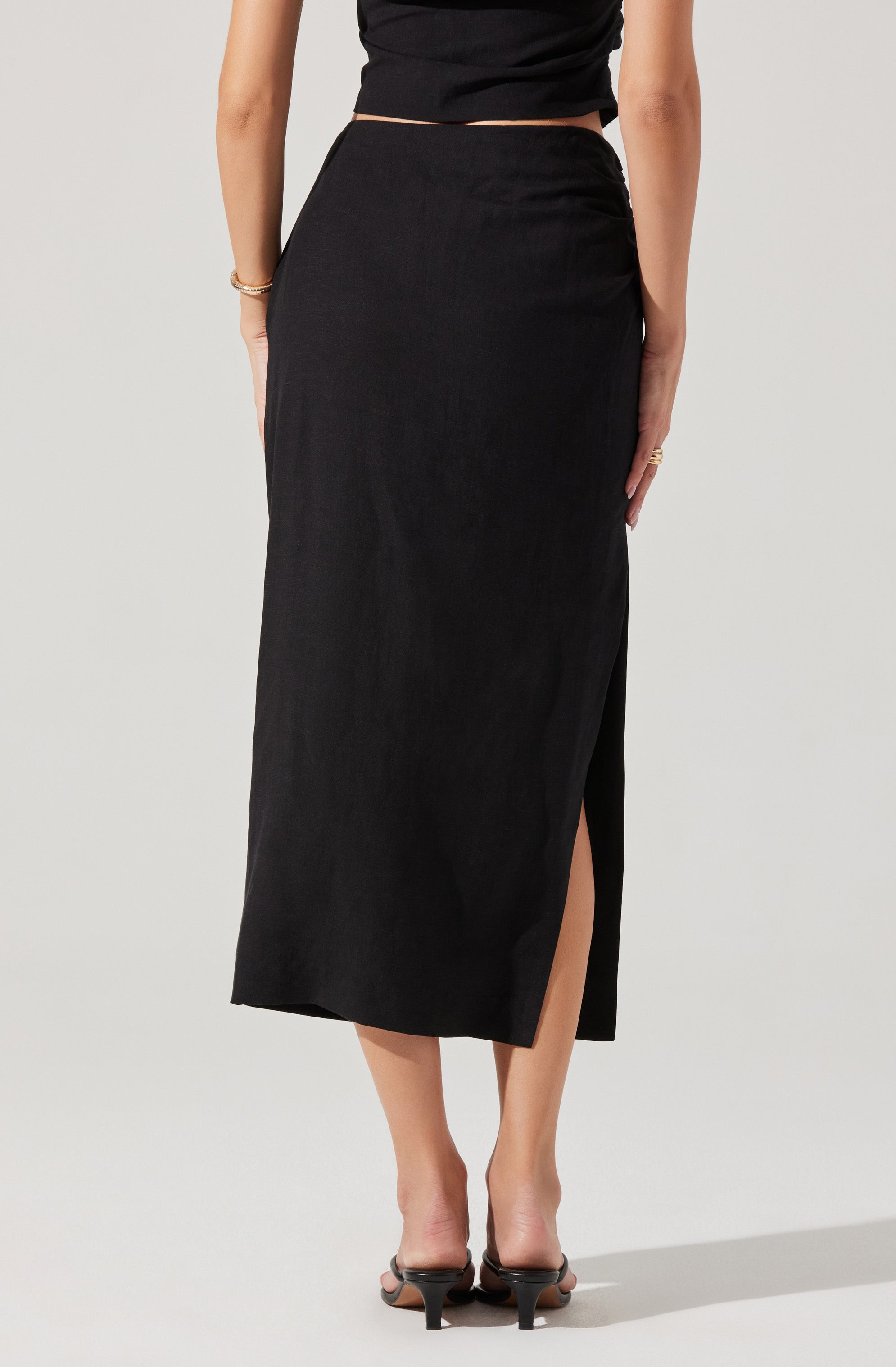 A woman is wearing a black, high-waisted, ankle-length skirt with a front slit on the left side. She pairs it with black open-toe heels and a black short-sleeved top. She accessorizes with a bracelet on her right wrist and a ring on her left hand.