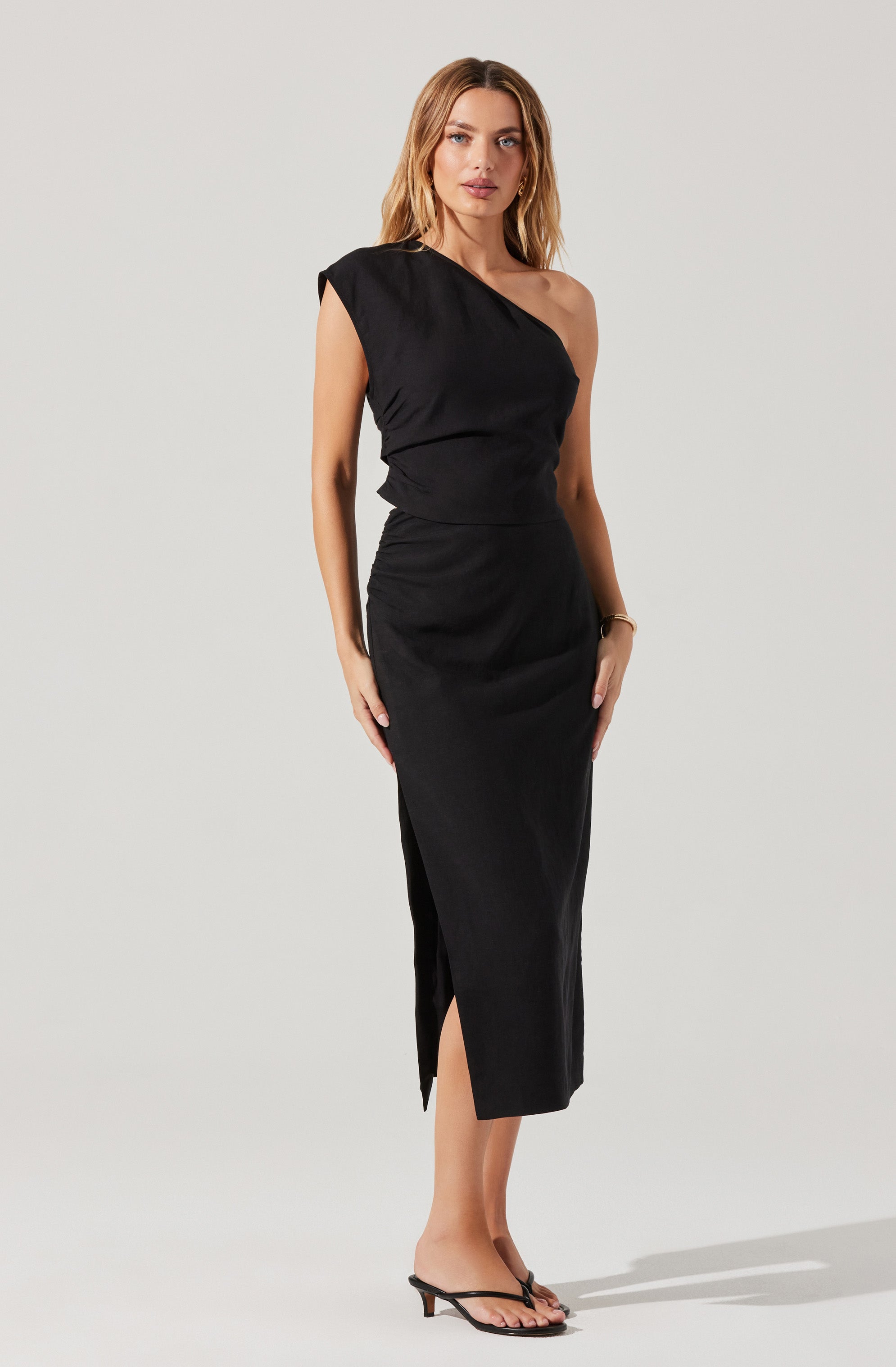 A woman is wearing a black dress with a one-shoulder design. The dress has a fitted bodice and a long, straight skirt that reaches mid-calf, with a small slit on the side. She is also wearing black, open-toed high-heeled sandals.