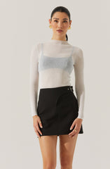 A woman is wearing a fitted, long-sleeve, ribbed sweater in a light, neutral color. The sweater appears semi-sheer with a subtle horizontal stripe pattern, revealing a hint of a light-colored top underneath. She is also wearing a high-waisted black mini skirt with a curved seam detail on the front and a decorative metal ring and grommet on the waistband. She accessorizes with large, round, gold earrings and rings on her fingers.