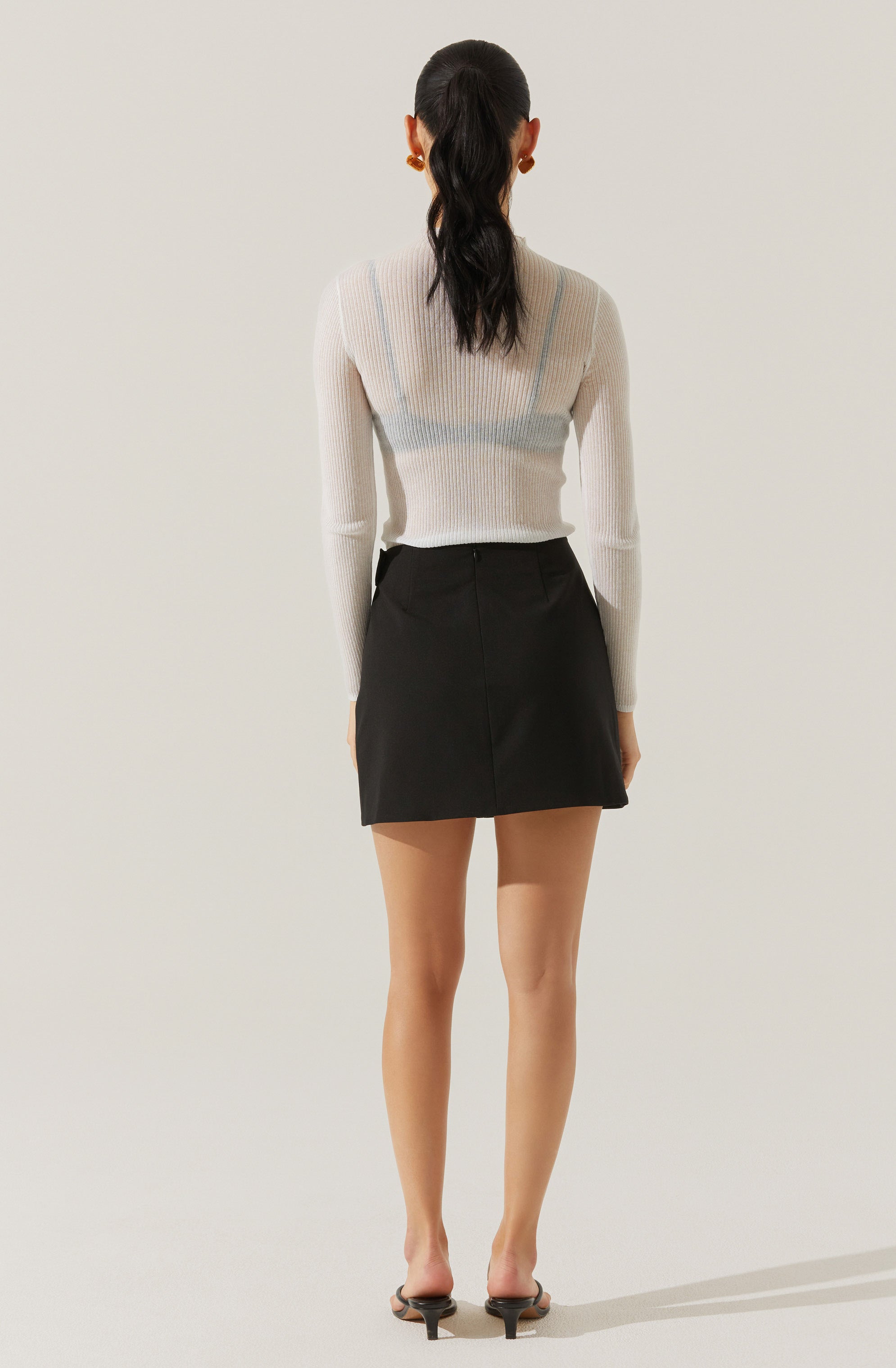 A woman is wearing a long-sleeved, semi-sheer, white ribbed sweater with a subtle vertical stripe pattern in light colors. She pairs it with a high-waisted, black A-line mini skirt. She has on black open-toe heeled sandals and is accessorized with large gold earrings. Her dark hair is styled in a low ponytail.