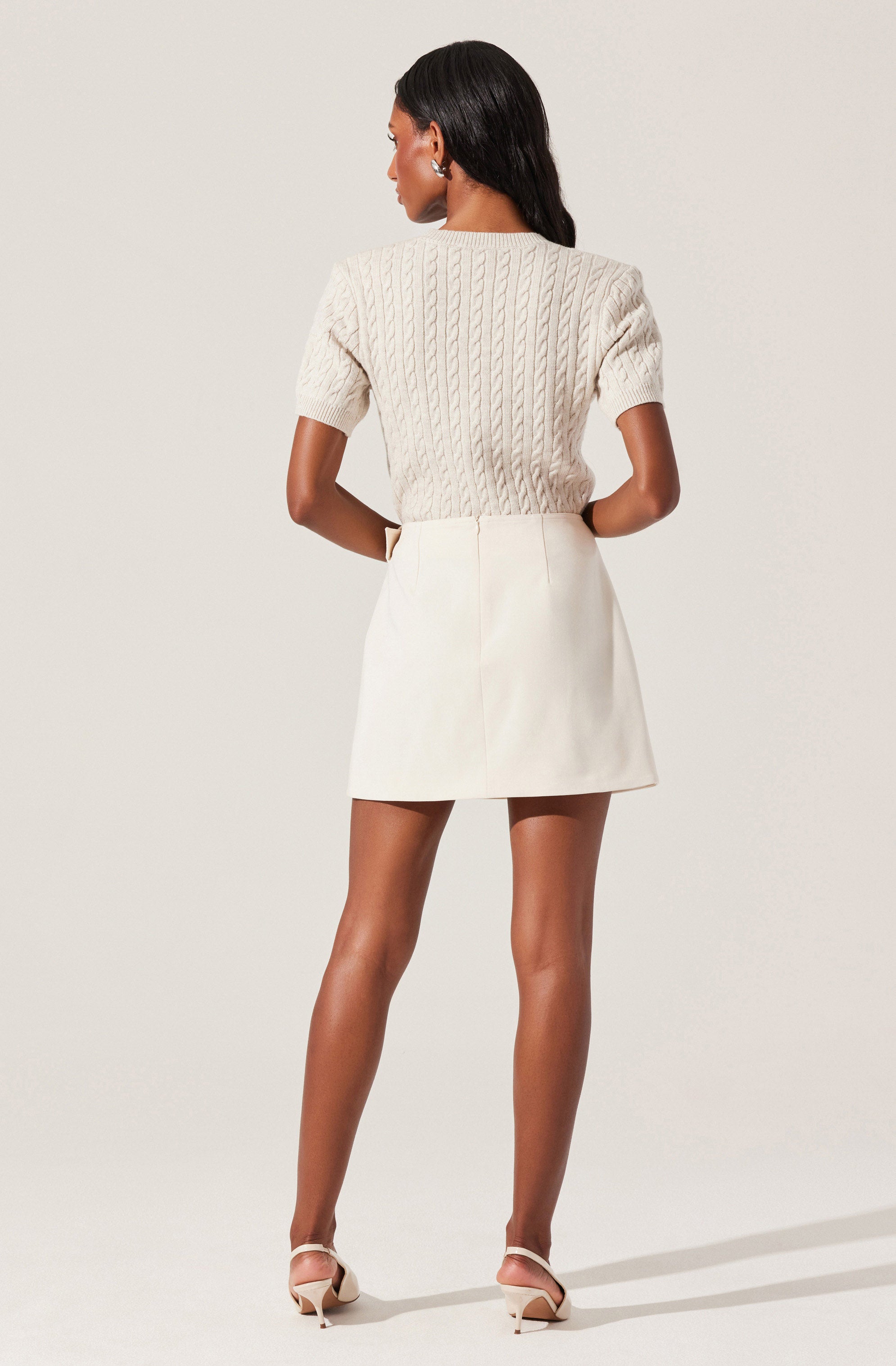 A woman is wearing a cream-colored, cable-knit short-sleeve sweater with a high neckline, paired with a matching high-waisted, A-line mini skirt. She is also wearing light-colored, open-toe high heels.