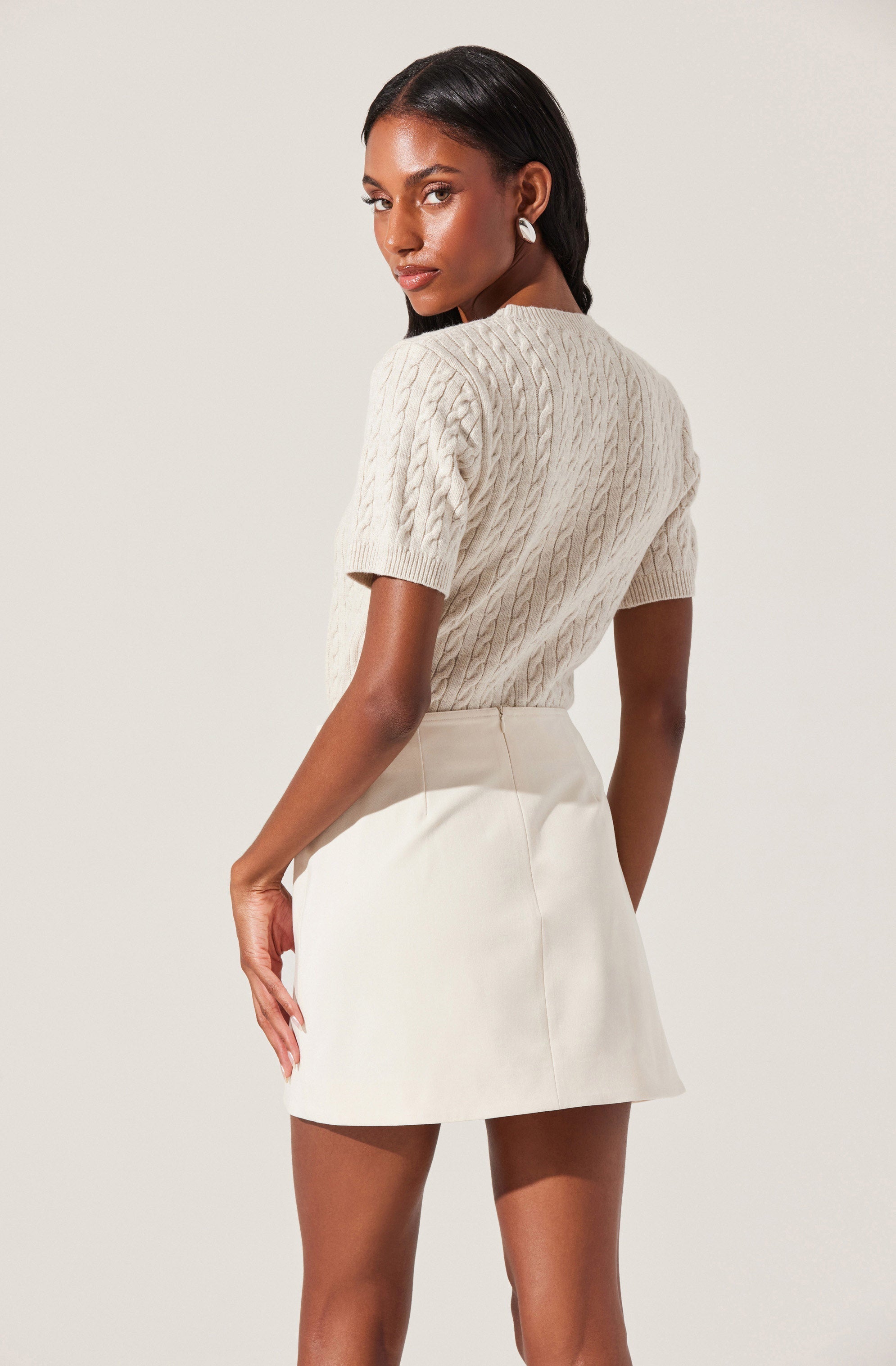 A woman is wearing a short-sleeved, cream-colored, cable-knit sweater and a high-waisted, off-white mini skirt.
