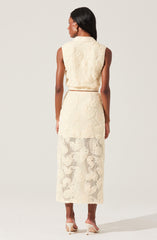 A woman is wearing a sleeveless, cream-colored textured two-piece outfit consisting of a cropped top with wide shoulders and a high-waisted, long skirt that extends to her ankles. The fabric has a subtle floral pattern with openwork details. She is also wearing nude high-heeled shoes.